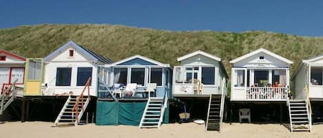 A row of beach houses with external stairs leading to the entrance, situated on a sandy beach with a grassy hill in the background.