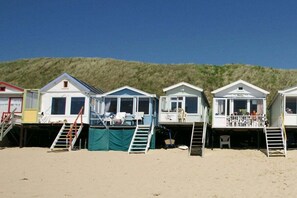 A row of beach houses with external stairs leading to the entrance, situated on a sandy beach with a grassy hill in the background. - Groepshuis Oase (12 people) (Zoutelande)