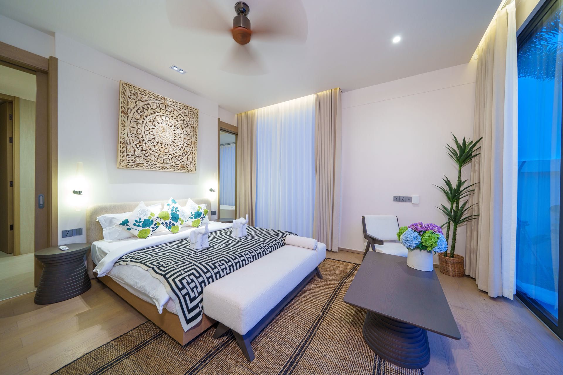 Una habitación de hotel moderna con una cama grande, mesitas de noche, un escritorio y un amplio ventanal con cortinas.
