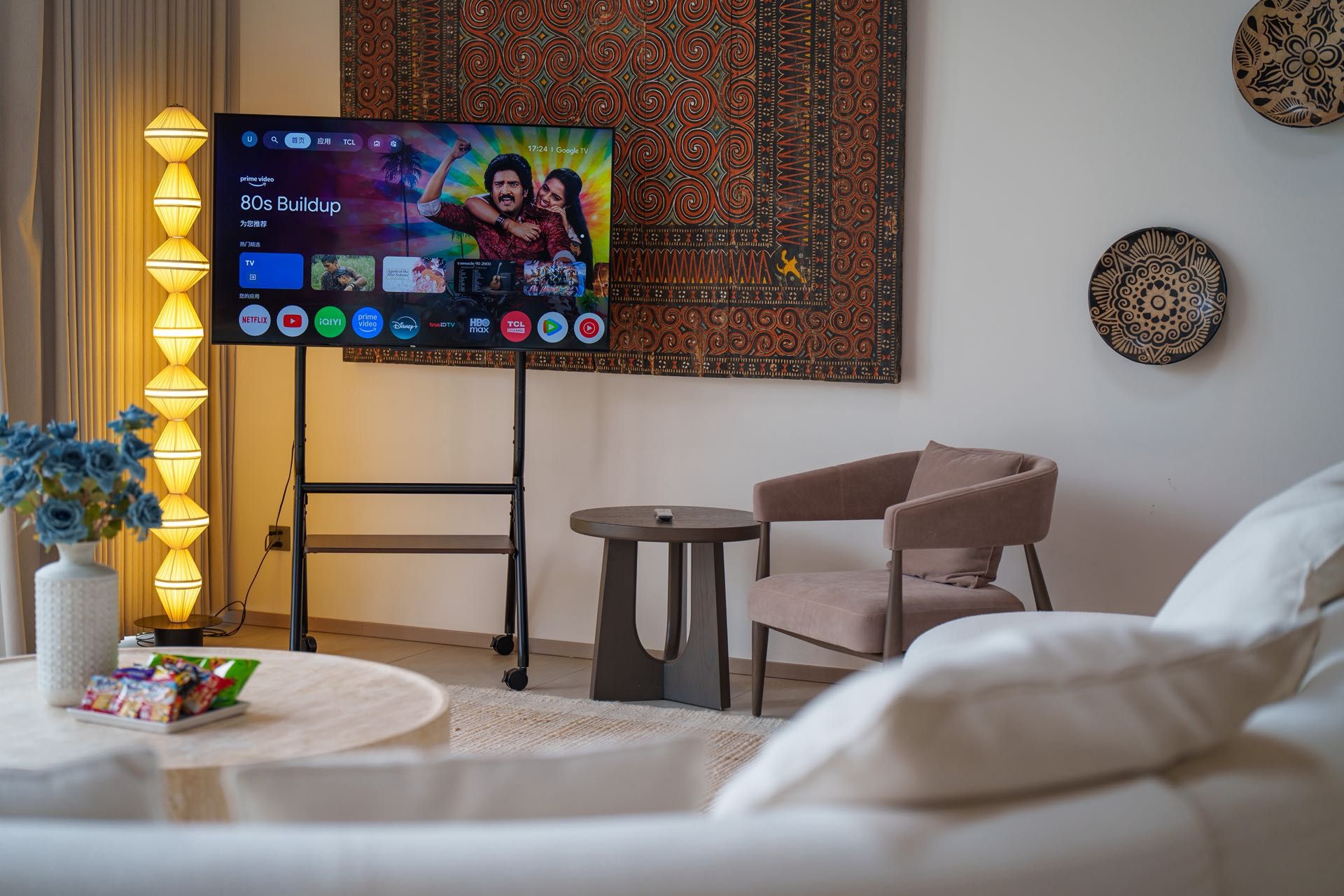 A hotel room with a flat-screen TV displaying a movie, a decorative wall tapestry, a small round table with a remote control, a cushioned chair, and a vase with flowers.