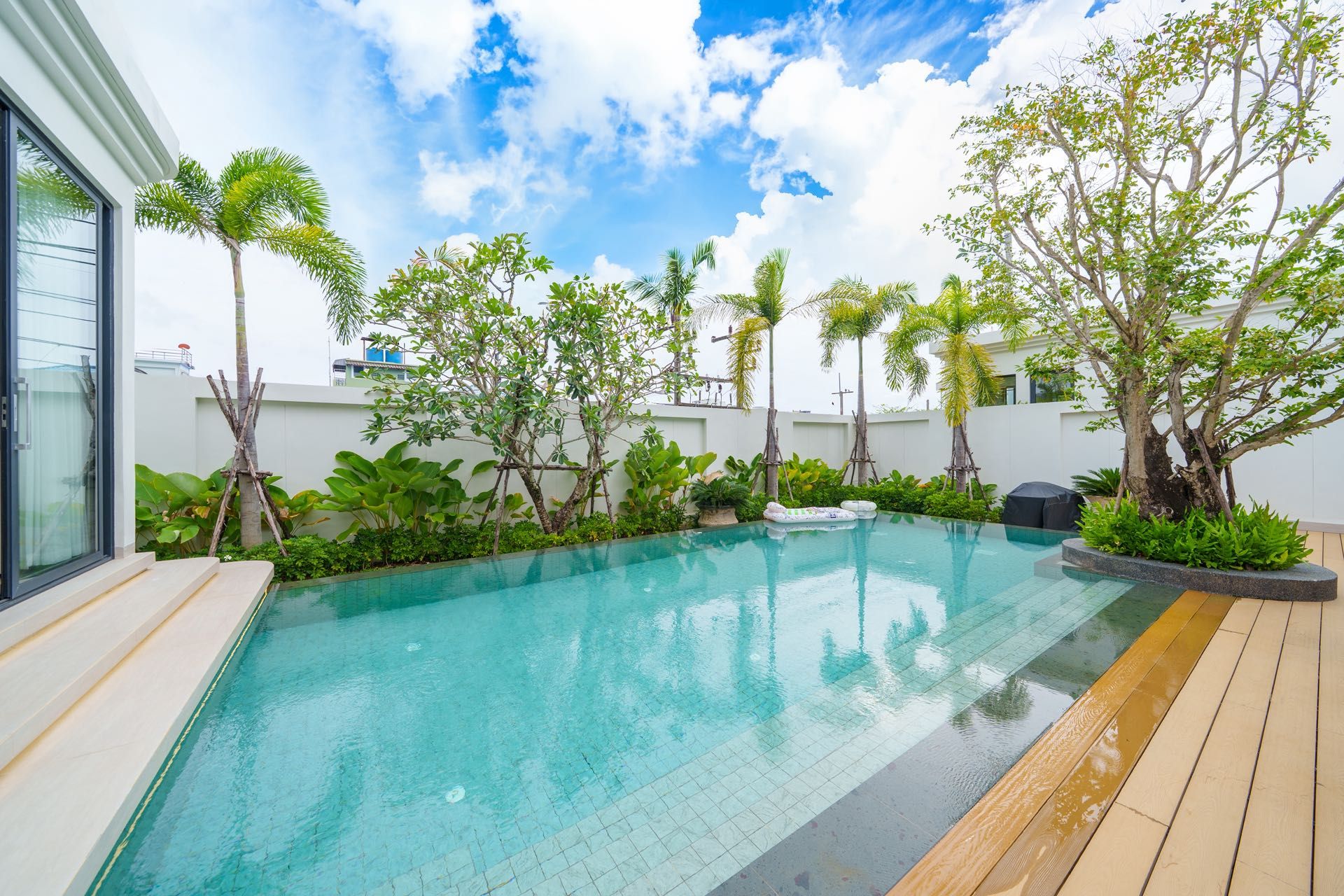 Una piscina con cubierta de madera, rodeada de plantas y árboles tropicales, bajo un cielo parcialmente nublado.