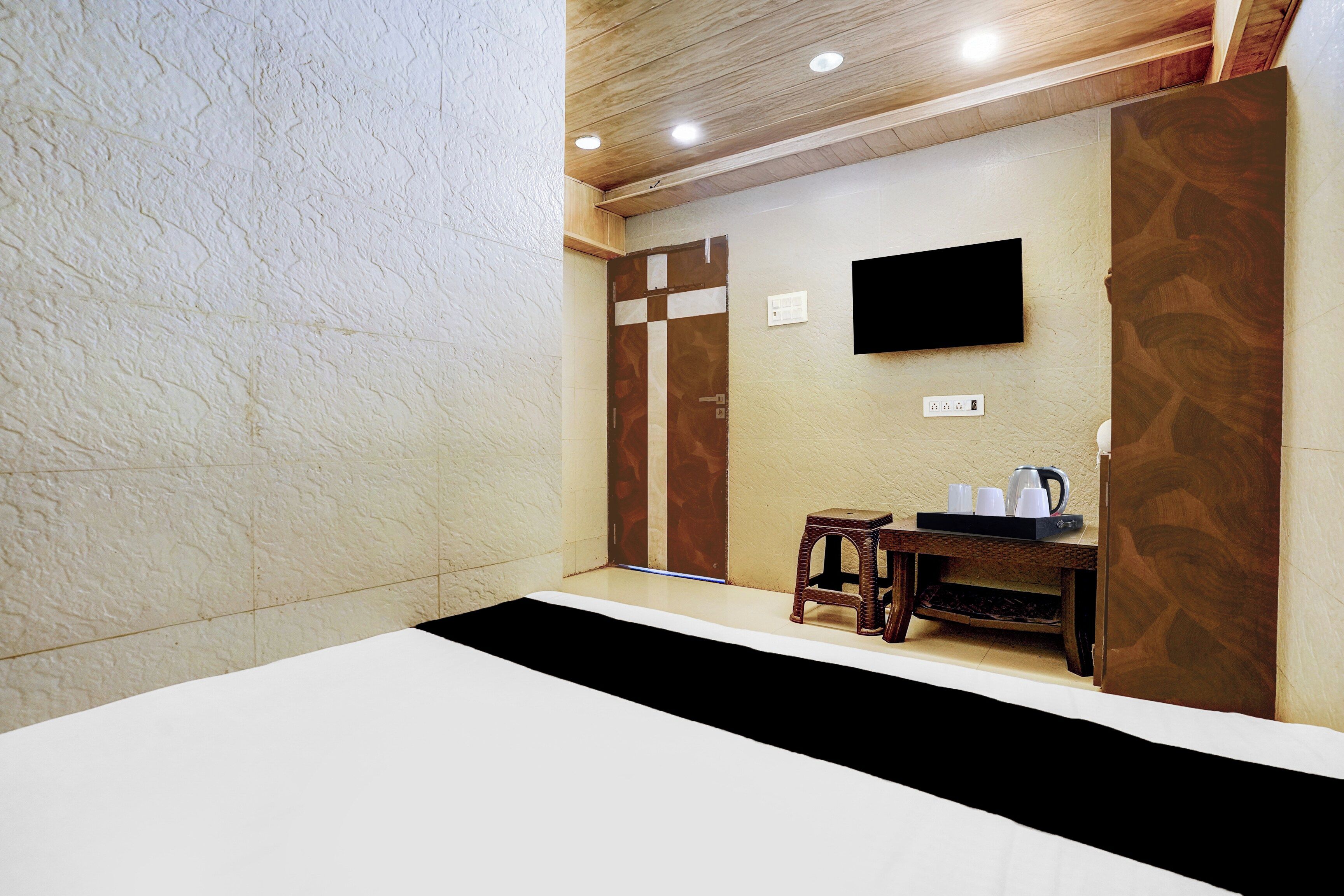Habitación de hotel con cama, una mesita con hervidor, televisor montado en la pared y puerta de madera.