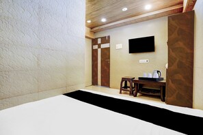 A hotel room with a bed, a small table with a kettle, a television mounted on the wall, and a wooden door.
