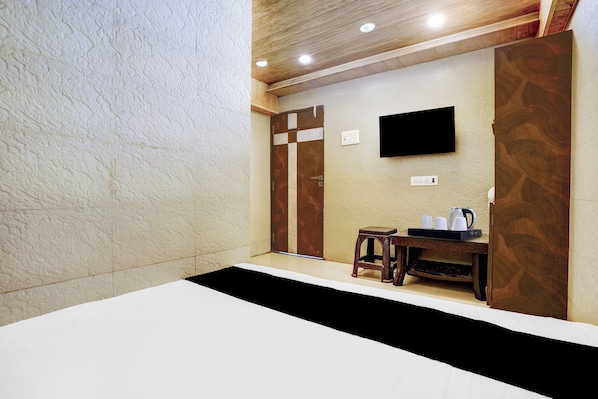A hotel room with a bed, a small table with a kettle, a television mounted on the wall, and a wooden door.
