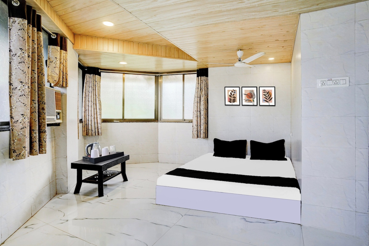 A bedroom with a bed, a table with cups and a kettle, a window with curtains, and wall-mounted artwork.