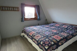 A bedroom with a large bed, a window with curtains, a radiator, and a wooden shelf. - Holiday apartment with 2 bedrooms (Julianadorp)