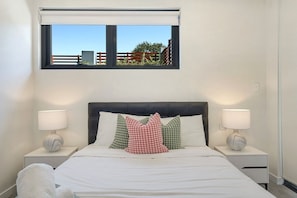 A bedroom with a bed, two bedside tables with lamps, a window with a view of a fence and greenery, and a headboard. - Elegant Urban Retreat in the Heart of Epping (Epping)
