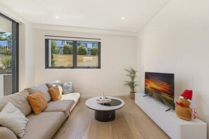 Una sala de estar moderna con un sofá, una mesa redonda de centro, un televisor de pantalla plana y una ventana con vistas a zonas verdes.