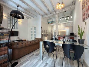 Un salon moderne avec un canapé en cuir marron, une table à manger en verre et des chaises noires, et un coin cuisine à l’arrière-plan.