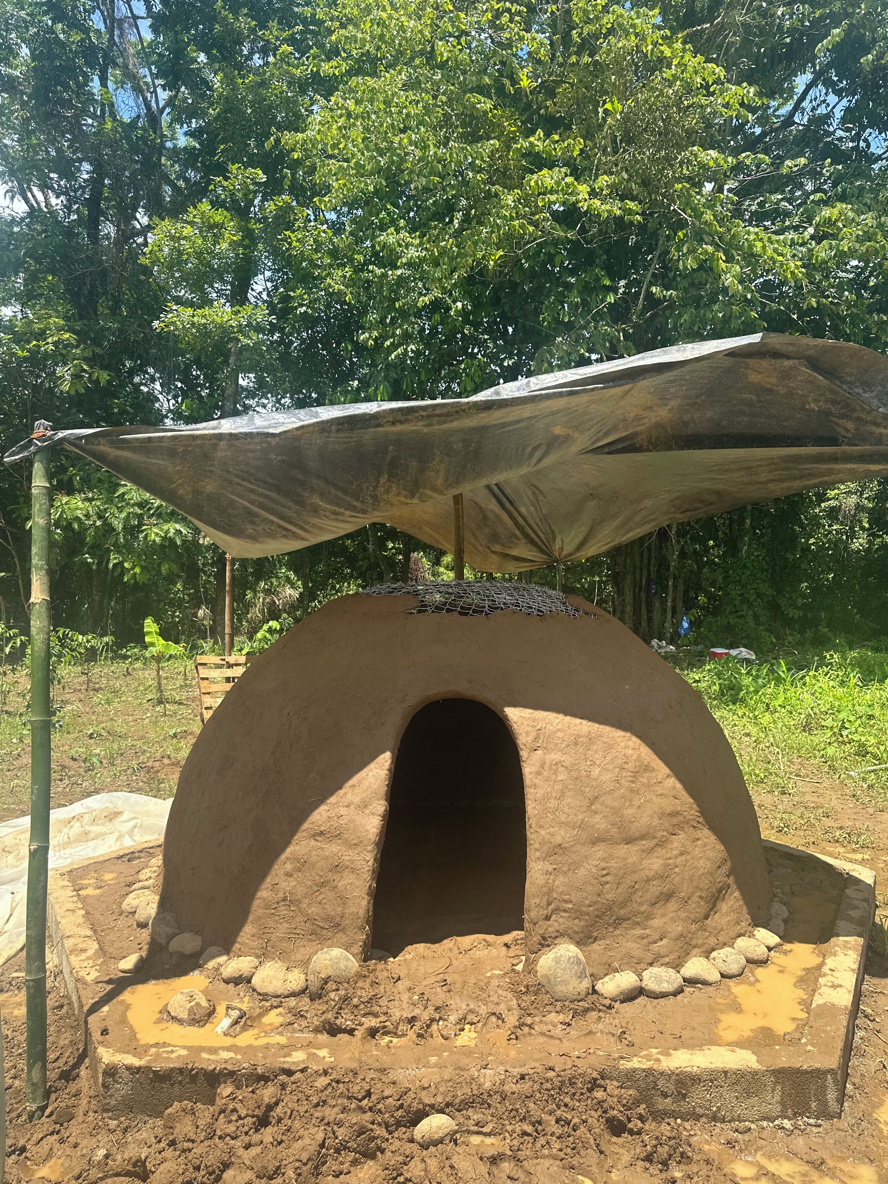 Un forno in argilla tradizionale con tetto di paglia, circondato da un’area sterrata con rocce e un palo di bambù.