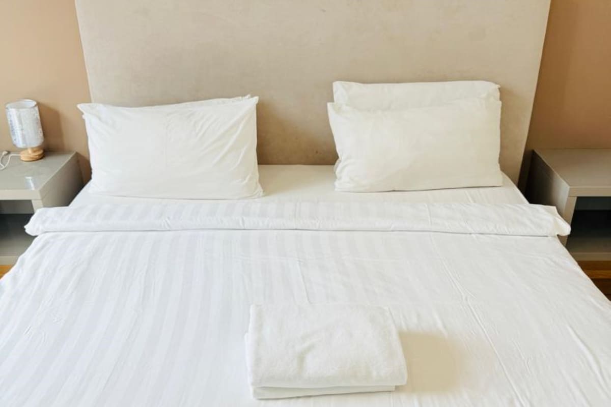 Una cama bien hecha con una toalla blanca doblada, dos almohadas blancas y una lámpara de noche sobre una mesita.