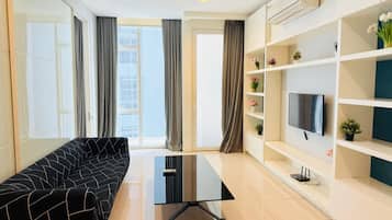 Una sala moderna con un sofá negro, una mesa de centro de vidrio, un televisor de pantalla plana sobre un estante blanco y un ventanal con cortinas.