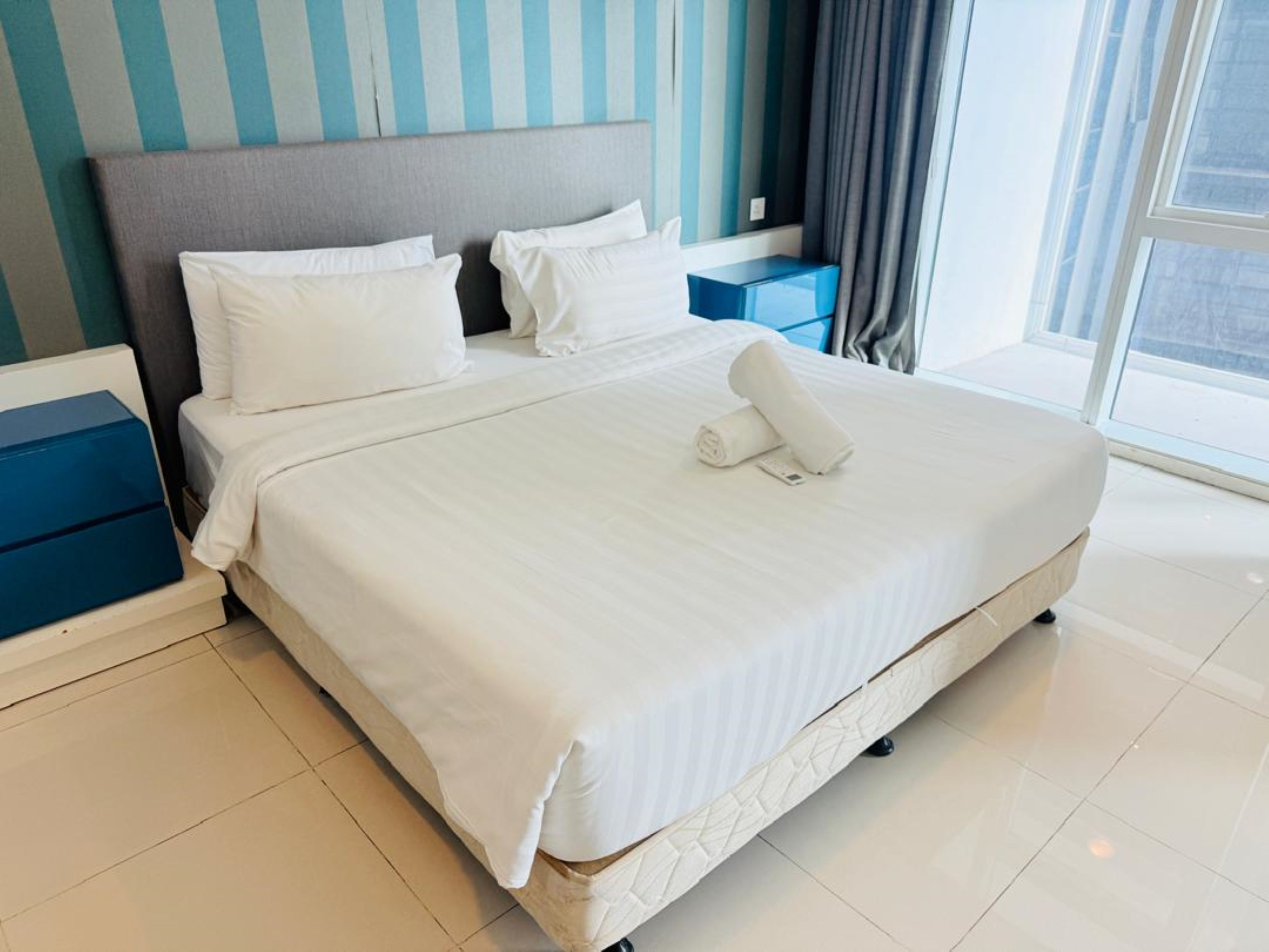 A neatly made bed with white linens, a grey headboard, and a blue bedside table with a blue box.