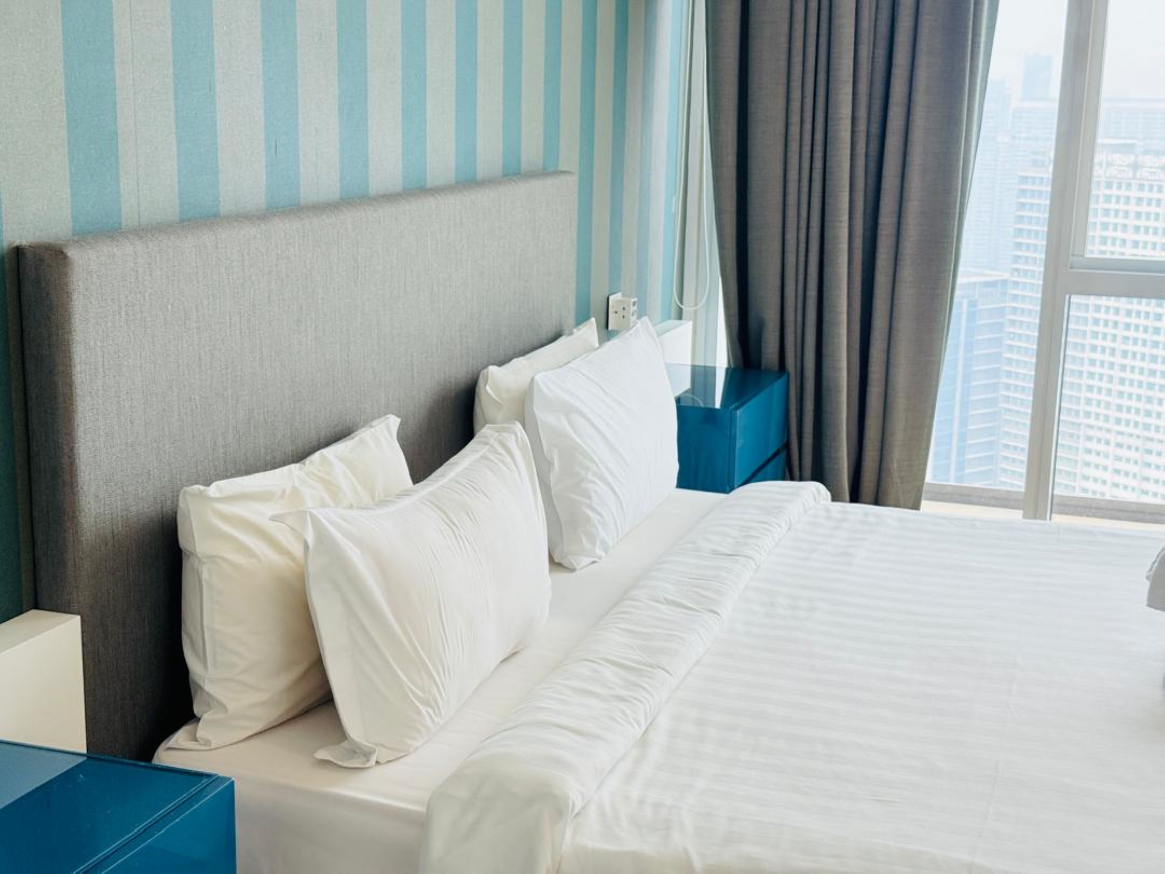 A hotel room with a bed, pillows, a nightstand, a blue bedside table, and a window with curtains.