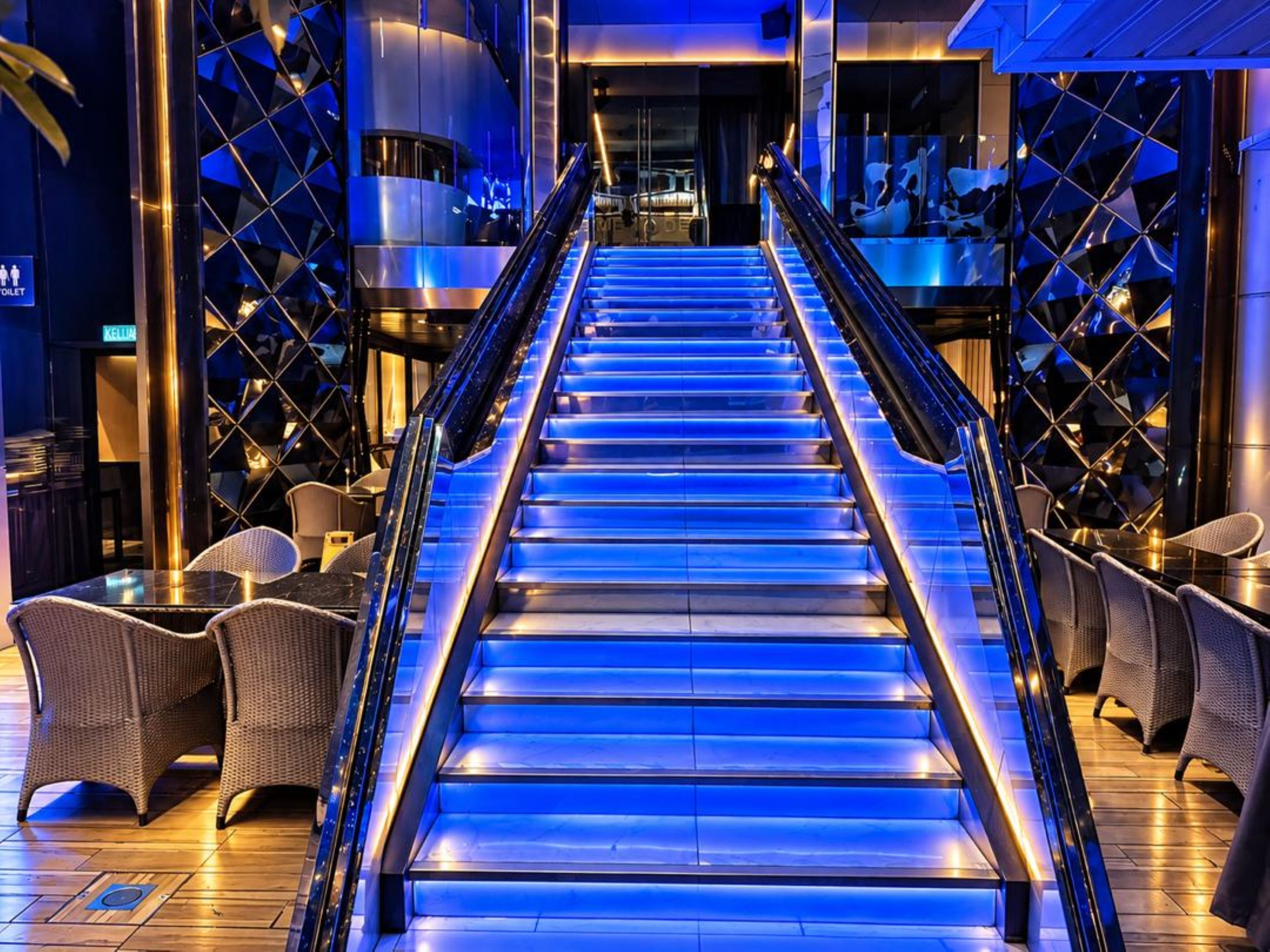 Una escalera con iluminación azul que lleva a una zona de bar con sillas de mimbre.