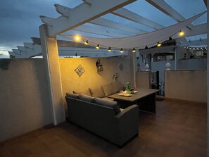 Ein überdachter Außenbereich mit einer Couch, einem Tisch, Lichterketten und dekorativen Wandmustern.