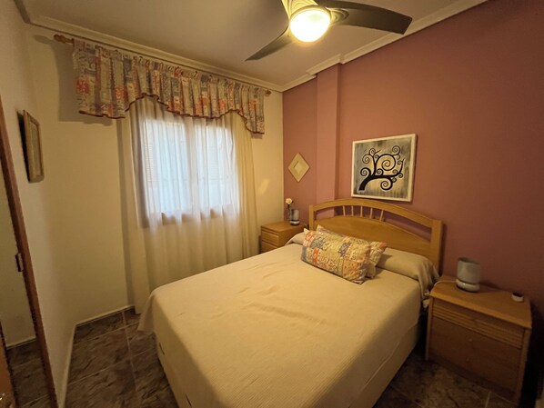 Ein Hotelzimmer mit Bett, Nachttisch, Wanduhr, Deckenventilator und einem Fenster mit Vorhängen.