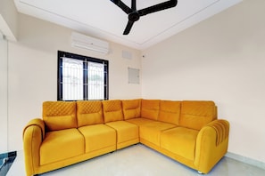 A room with a yellow sectional sofa, a ceiling fan, an air conditioning unit, a window with metal bars, and a whiteboard on the wall. - Super Hotel O Vilankurichi (Coimbatore)