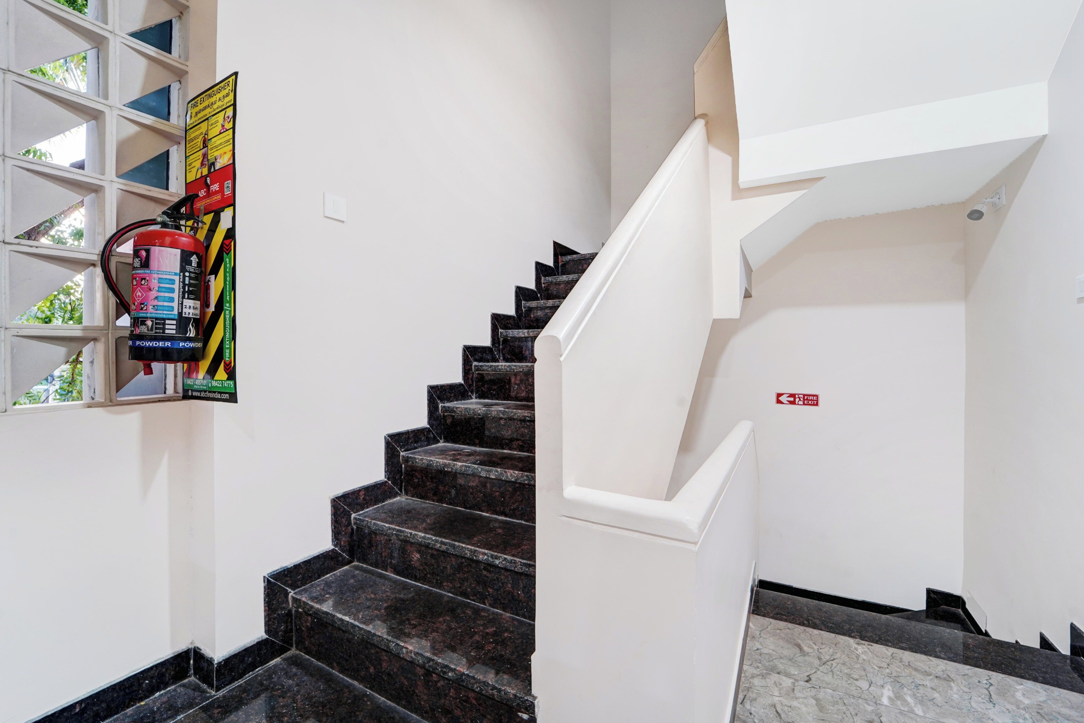 Eine Treppe mit schwarzen Marmorstufen, die zu einer Zwischenebene führt, ein an der Wand befestigter Feuerlöscher und ein Feuerausgangsschild.