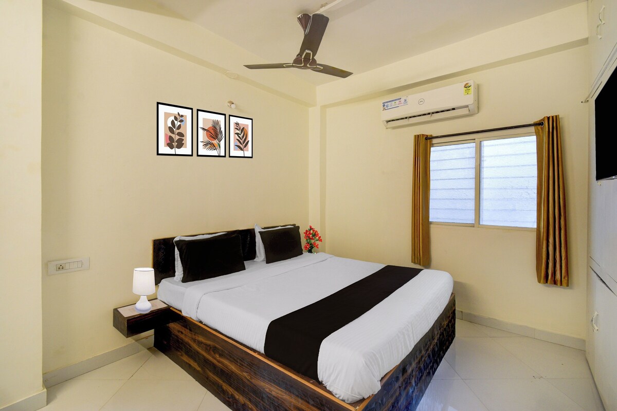 Quarto de hotel com cama, mesa de cabeceira, luminária, aparelho de ar condicionado, janela com cortinas e obra de arte na parede.