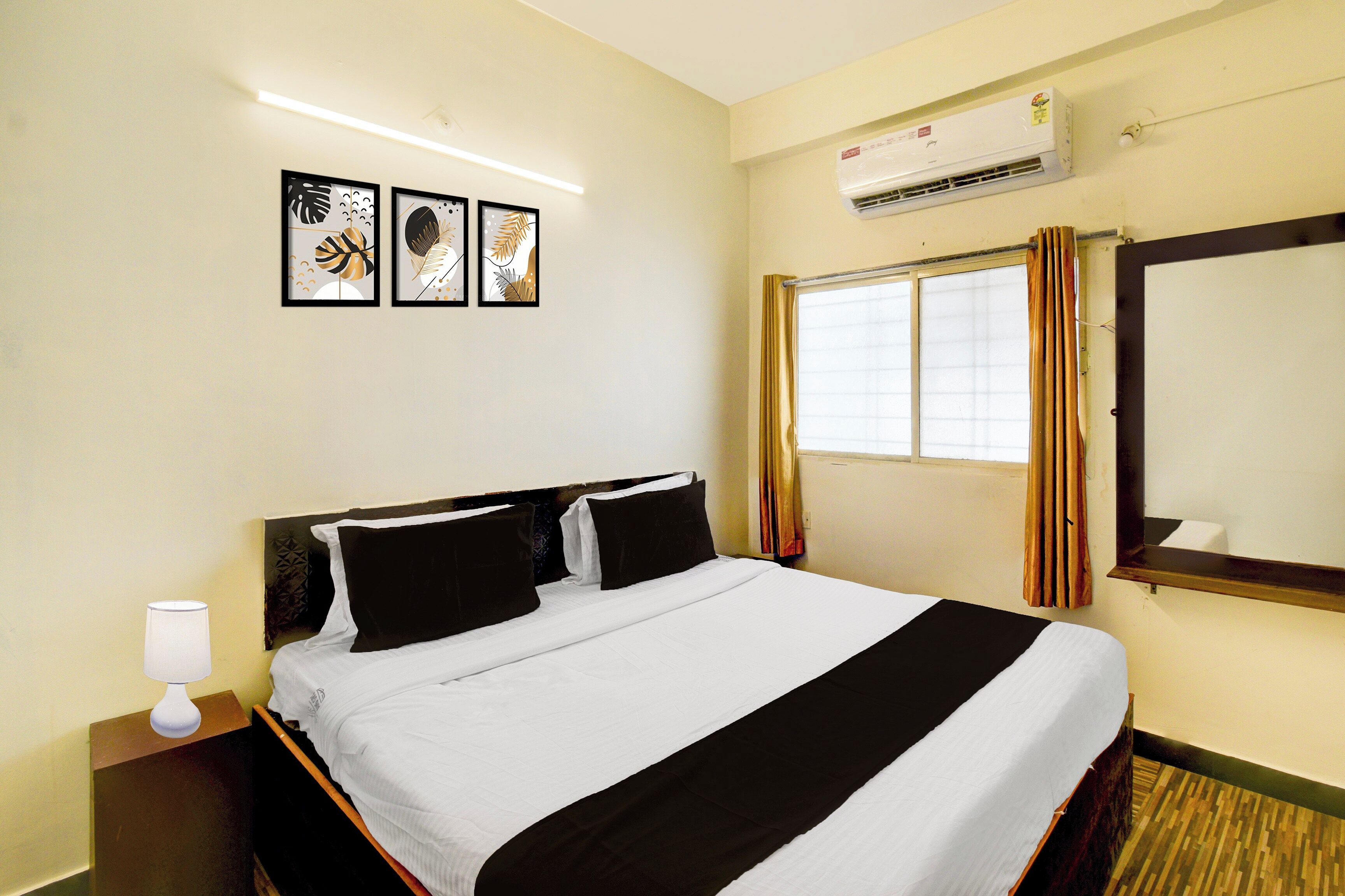 A hotel room with a bed, a nightstand, a lamp, an air conditioner, a window with curtains, and three framed pictures on the wall.