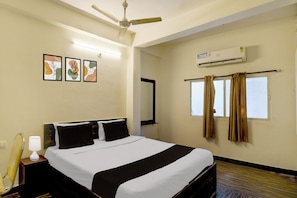 Quarto de hotel com cama, ventilador de teto, ar condicionado, janela com cortinas e três quadros na parede.