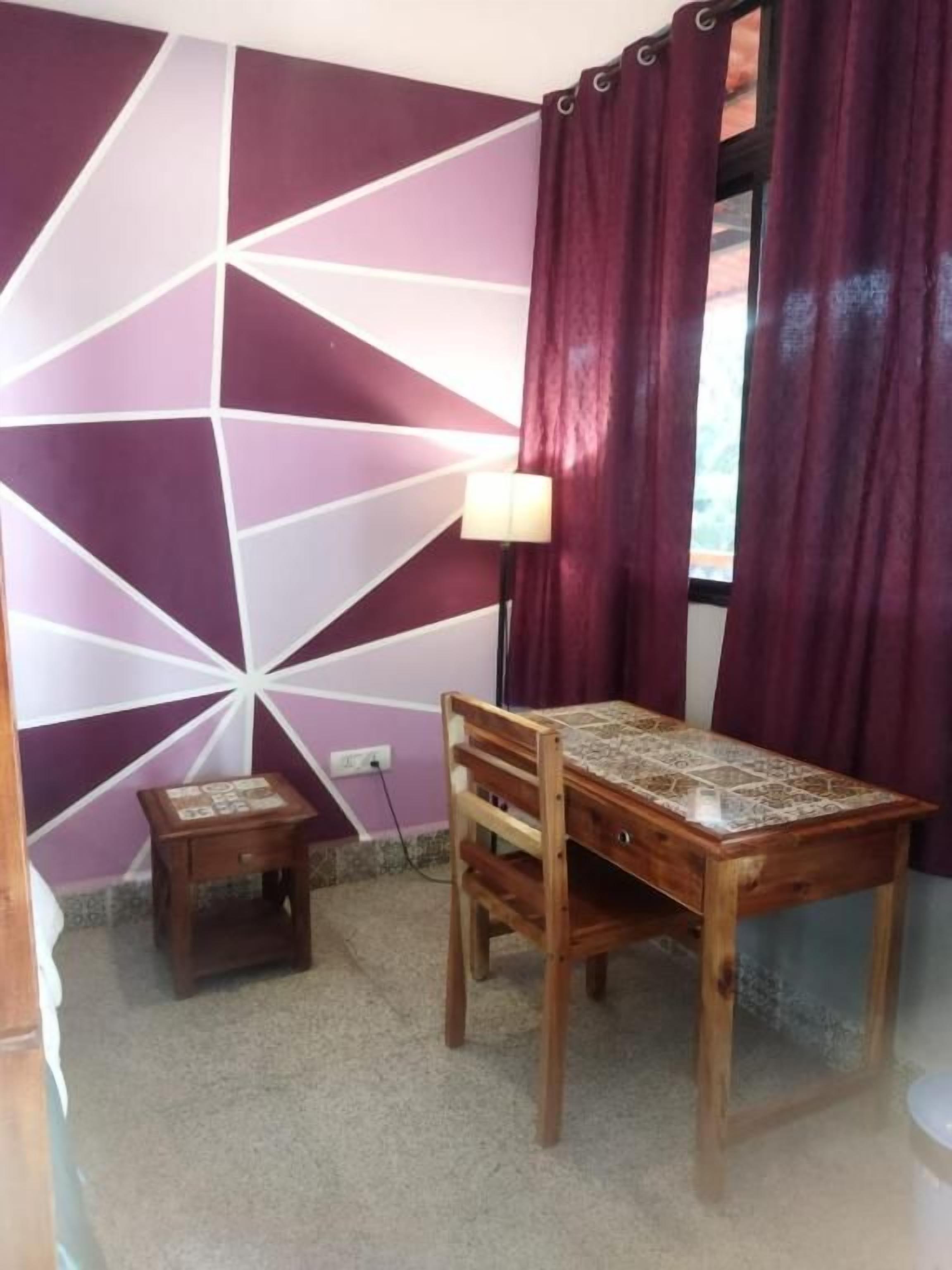 Una habitación con una mesa de madera, un taburete pequeño, una lámpara y una pared con diseño geométrico.