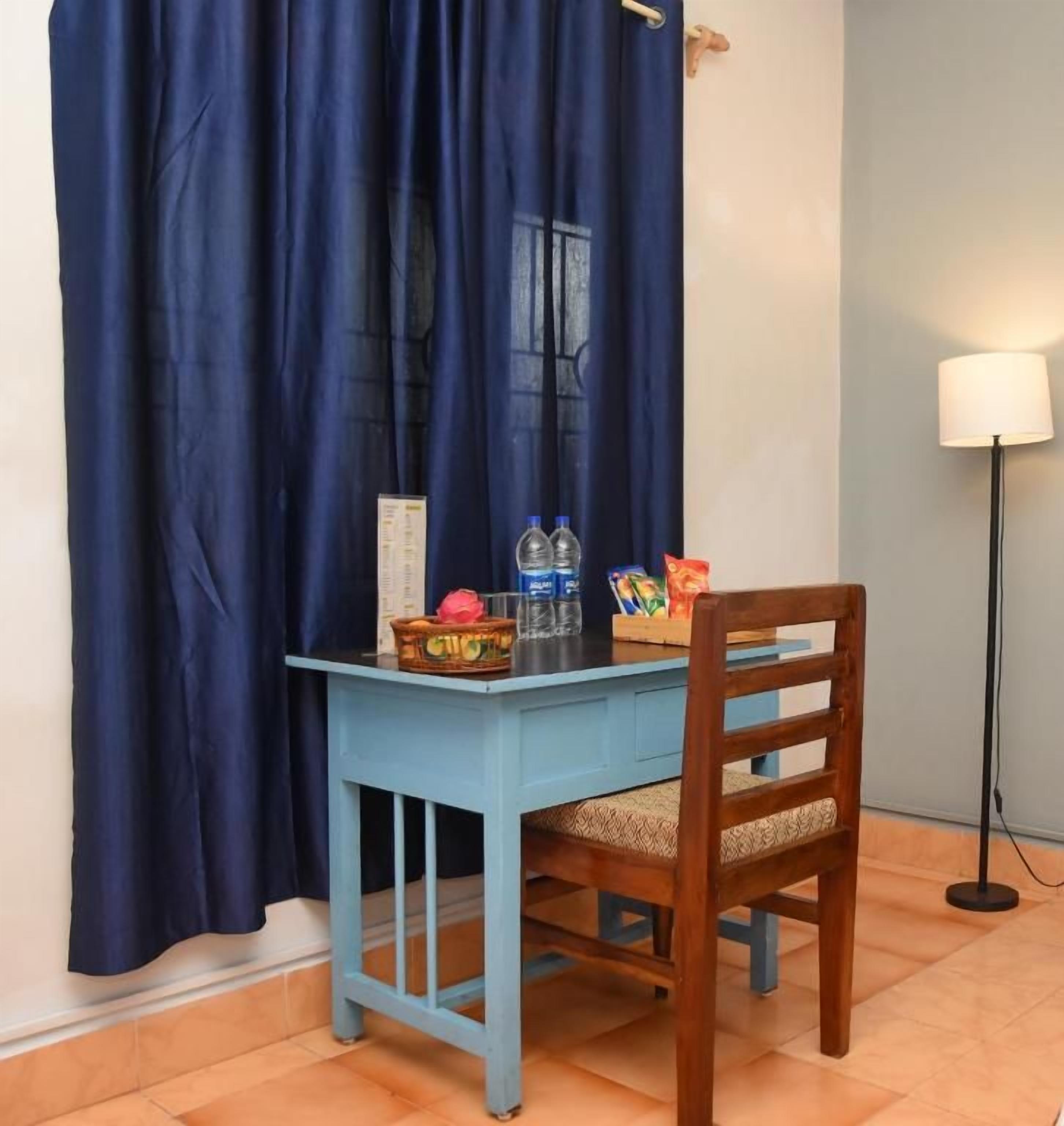 Una habitació amb taula i cadira blava, una làmpada i una finestra amb cortines.