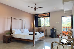 A four-poster bed with a ceiling fan, a small table with a chair, a refrigerator, and a window with a view of greenery. - Bens Inn Ashwem Beach (Ashvem)