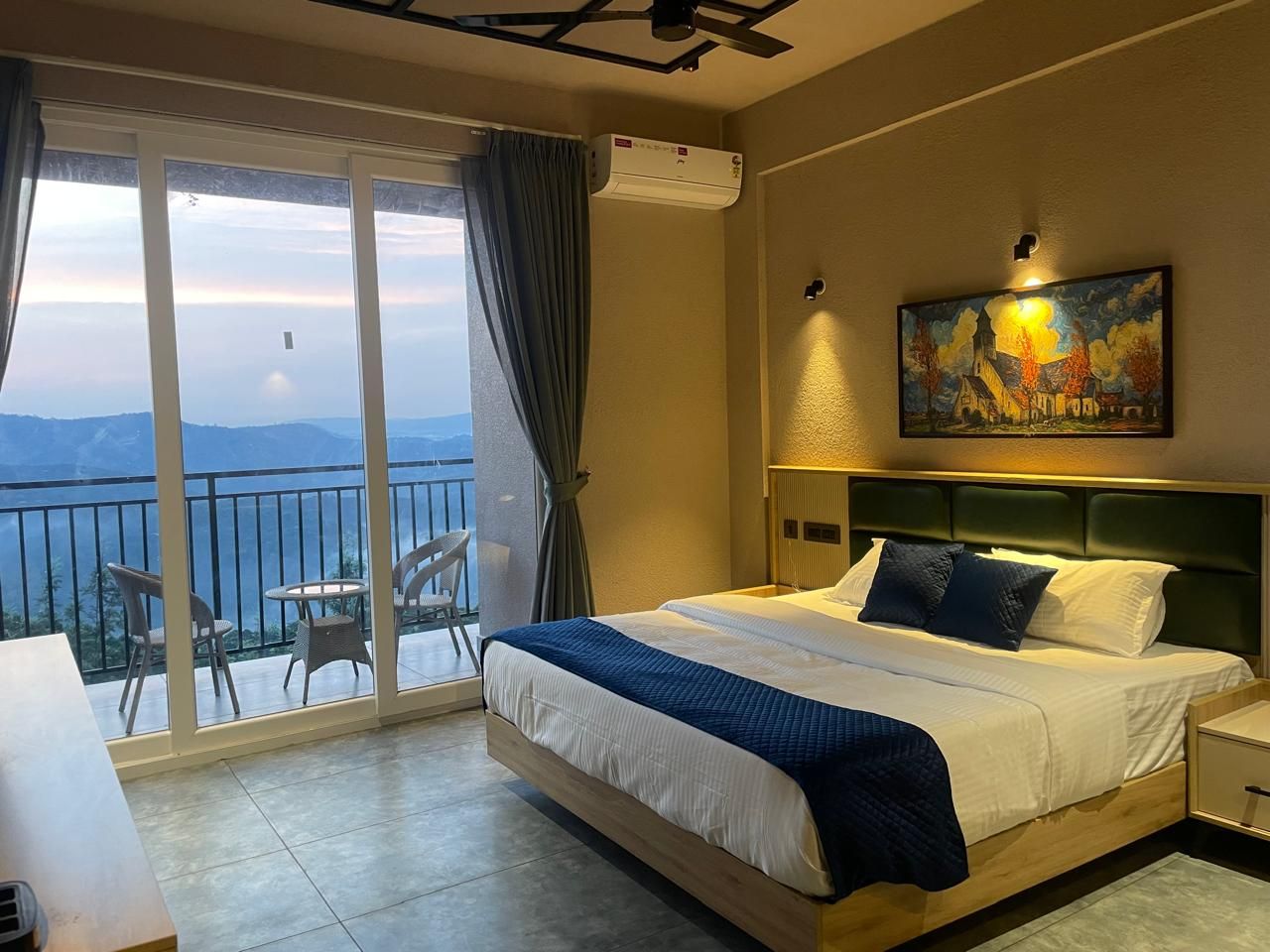 Une chambre d’hôtel avec un lit, un tableau, un balcon avec des chaises et une vue sur les montagnes.