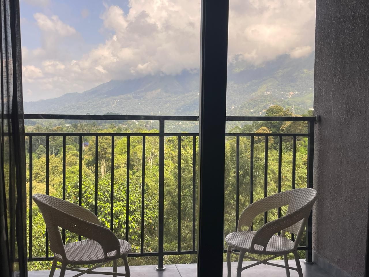 Un balcón con dos sillas de mimbre, baranda metálica y vista a un paisaje montañoso.