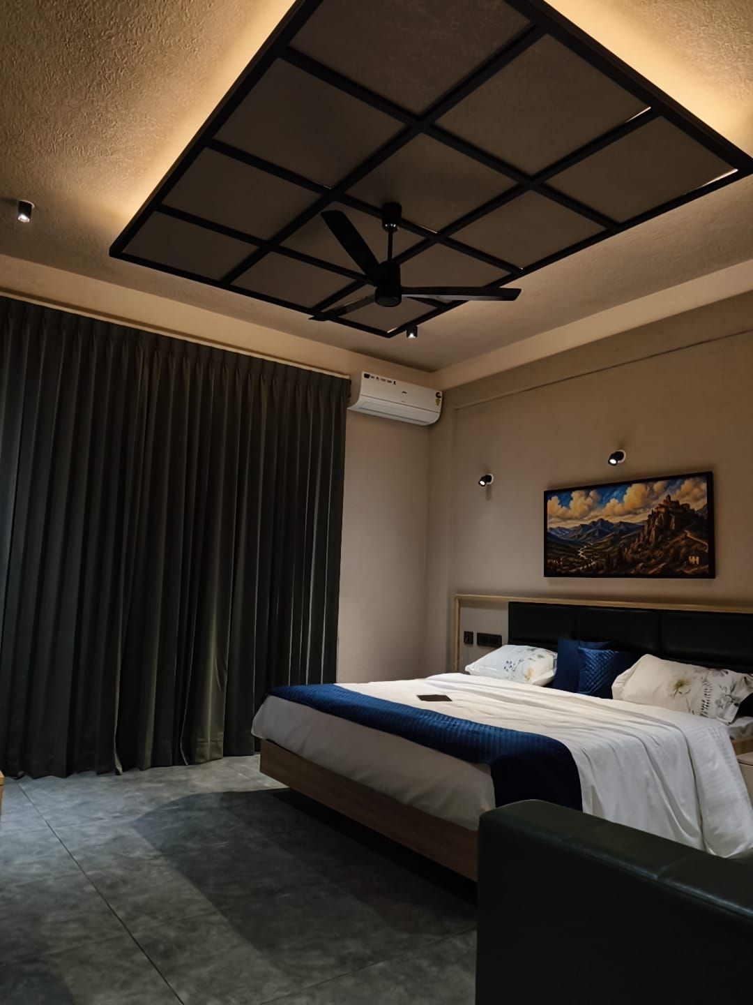 Een hotelkamer met een bed, een plafondventilator, een airconditioner, een schilderij aan de muur en een raam met gordijnen.