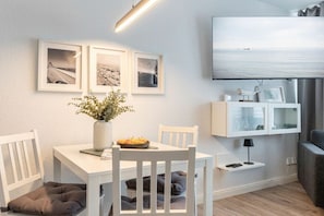 Ein modernes Wohnzimmer mit einem weißen Esstisch, Stühlen, einer Blumenvase und einem flachbildfernseher an der Wand.