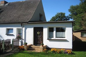 A single-story house with a sloped roof, a front door with a wreath, a small porch, and a garden with flowers. - Holiday apartment with 2 bedrooms (Schaprode)