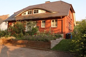 A two-story brick house with a sloped roof, a front garden with various plants, and a visible trash bin. - Apartment on the west side (Gager)