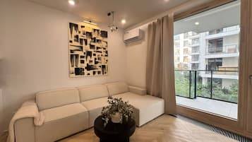 A modern living room with a beige sofa, a black coffee table with a plant, a geometric wall art piece, and a view of a residential building through a large window.