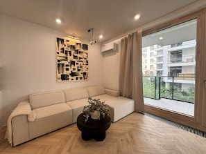A modern living room with a beige sofa, a black coffee table with a plant, a geometric wall art piece, and a view of a residential building through a large window. - Chmielna IV P&O Apartments (Warszawa)