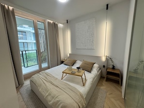 A modern bedroom with a large bed, a bedside table, a nightstand, a window with curtains, and a view of greenery outside. - Chmielna IV P&O Apartments (Warszawa)
