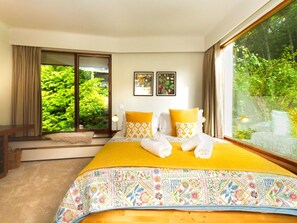 A bedroom with a large bed, two pillows, a bedside table, a window with a view of greenery, and two framed pictures on the wall.