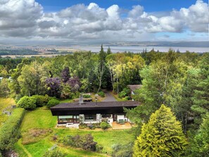 A modern house with a large glass window, surrounded by lush greenery and a view of a lake.