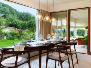 Ein Essbereich mit einem großen Tisch, gedeckt für sechs Personen, umgeben von Stühlen, mit Blick auf einen üppigen Garten und einer Glastür zum Außenbereich.