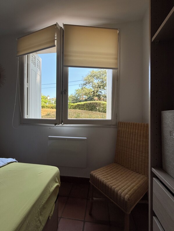 A small room with a bed, a chair, a radiator, and a window with a view of greenery and a building. - New Listing (Royan)