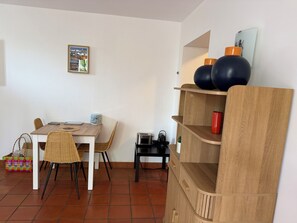 A dining area with a wooden table, wicker chairs, a small side table with a toaster, and a decorative shelf with two vases. - New Listing (Royan)
