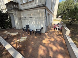 An outdoor patio area with a stone floor, two lounge chairs, a small table with a bottle and glasses, and a white table with chairs. - New Listing (Royan)