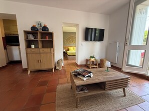 A living room with a wooden coffee table, a bookshelf, a television, and a view through a glass door. - New Listing (Royan)