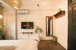Ett hotellrum med en säng, en väggmonterad tv som visar ”NETFLIX”, en krukväxt, en hylla i trä och en dörr med en spegel.