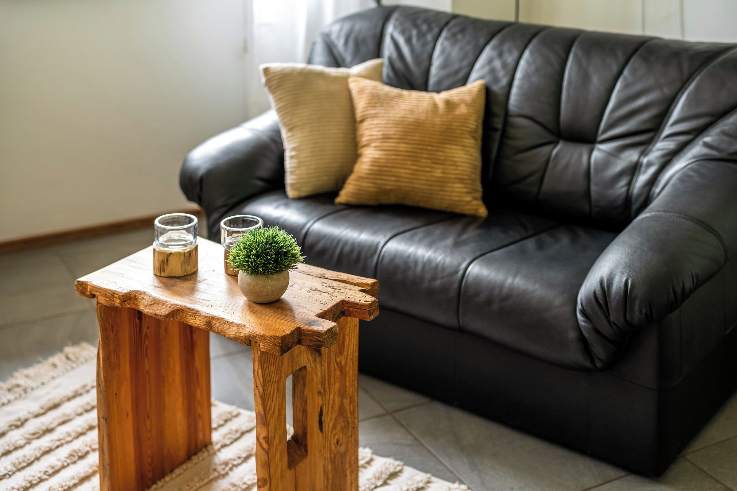 Een houten salontafel met twee glazen en een kleine plant, voor een zwarte leren bank met een beige kussen.