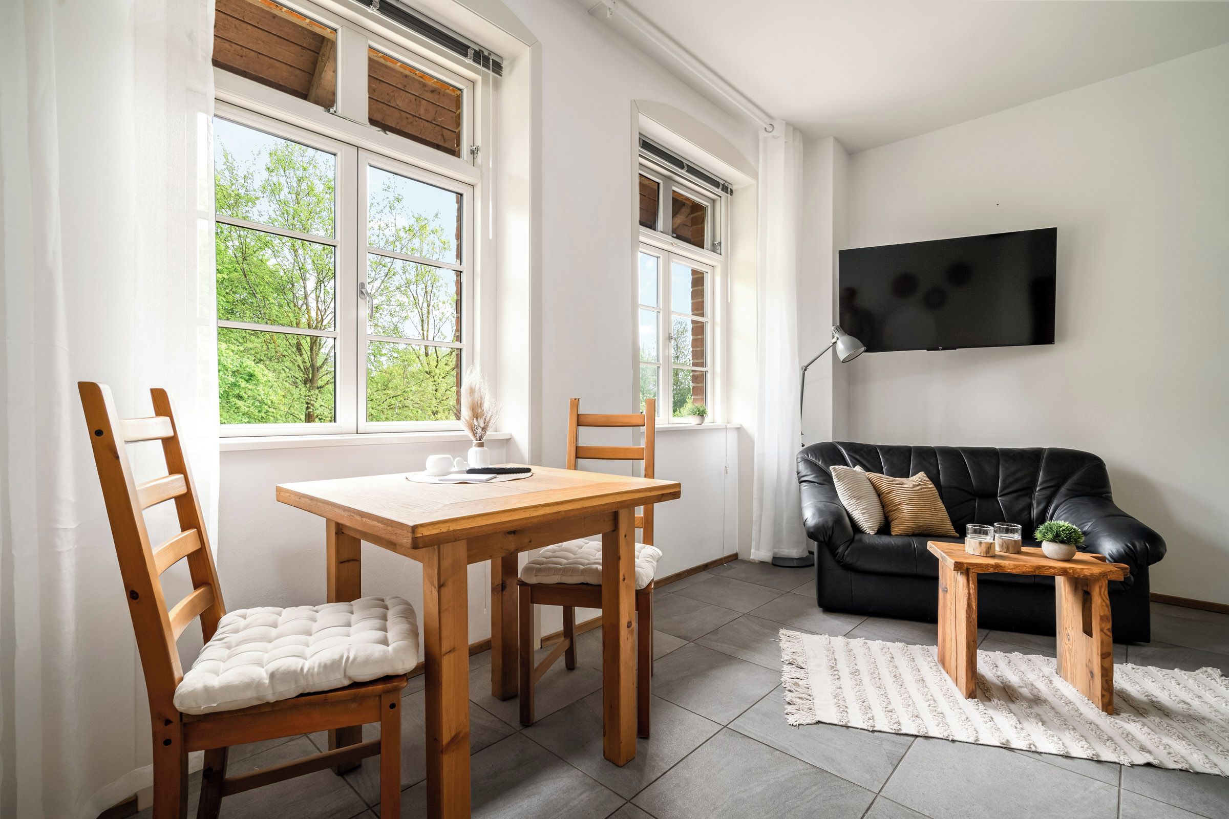 Ein modernes Wohnzimmer mit einem schwarzen Ledersofa, Holzstühlen, einem kleinen Holztisch und einem an der Wand befestigten Fernseher.
