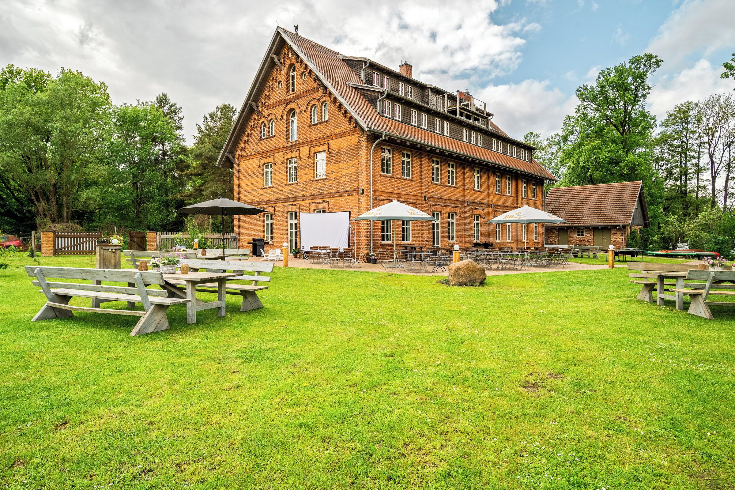 A large brick building with multiple windows and a sloped roof, surrounded by a grassy area with picnic tables and umbrellas.