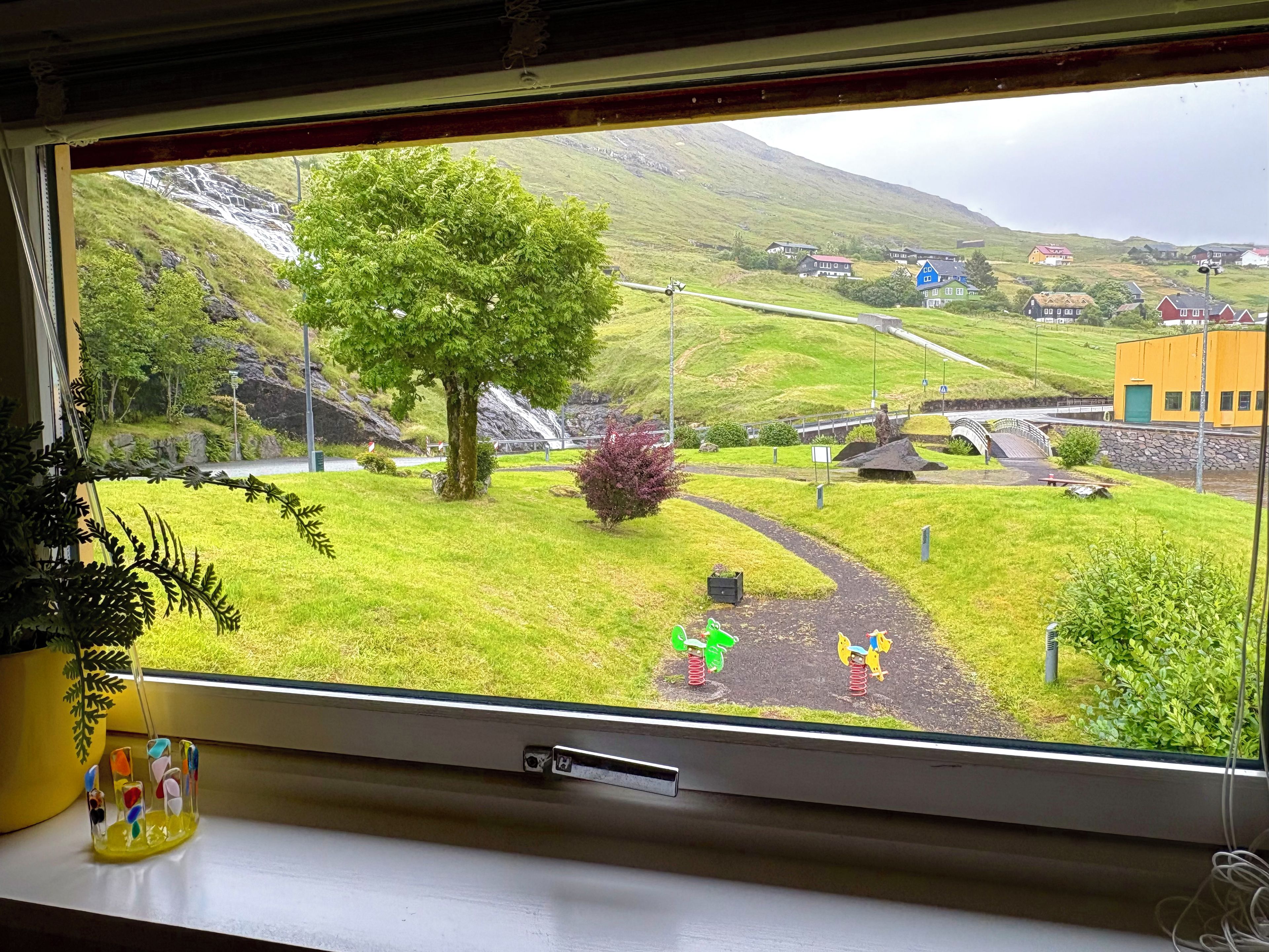 A window view of a green landscape with a waterfall, a tree, and a yellow building.